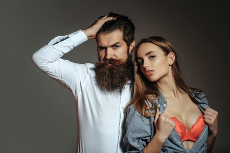 Young sexy couple of bearded handsome man hipster with beard and moustache and pretty girl or beautiful woman in red sexi bra in unbutton shirt on grey wallの写真素材