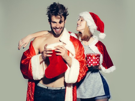 happy young couple of pretty cute girl or woman and handsome bearded man with bare muscular chest in santa claus coat and hat with new year sock or stocking and present or gift box on grey backdropの写真素材