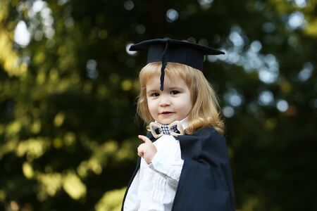 Little boy child in black academic gown and squared school hat and bow tie with blonde hair on smiling face standing outdoor on green natural backgroundの写真素材
