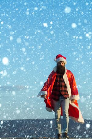 young handsome bearded bad santa claus man with long beard in checkered shirt jeans and red new year hat in christmas or xmas coat outdoor on blue sky background, copy spaceの写真素材