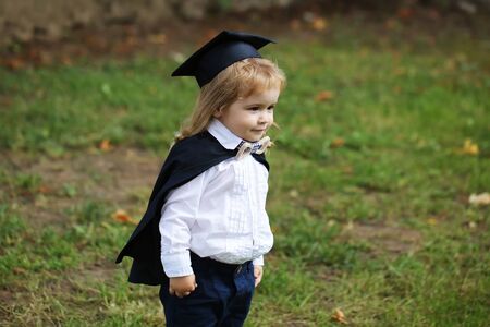 Little boy child in black academic gown and squared school hat with blonde hair standing on green grass outdoor on natural backgroundの写真素材