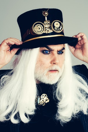 Senior serious bearded man or watchmaker with blue color lenses in eyes and white beard hair in black hat with watch mechanical metallic gears and cogwheels on grey wallの写真素材