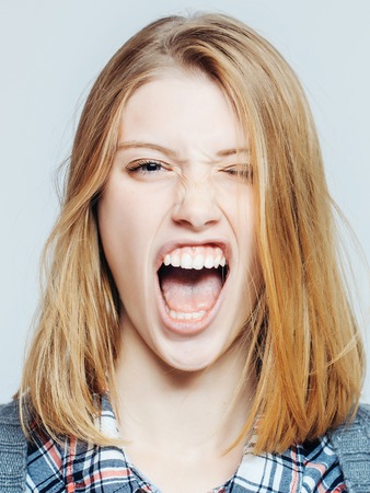 Angry pretty girl or cute beautiful woman young female model with blonde hair in plaid shirt and knitted coat grimaces and shows teeth on grey backgroundの写真素材
