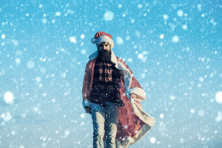 young handsome bearded bad santa claus man with long beard in checkered shirt jeans and red new year hat in christmas or xmas coat outdoor on blue sky backgroundの写真素材