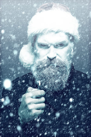 Closeup portrait of one senior frozen new year man with painted face in white and long beard in christmas santa red hat holding cigarette lighter in studio on black background, vertical pictureの写真素材
