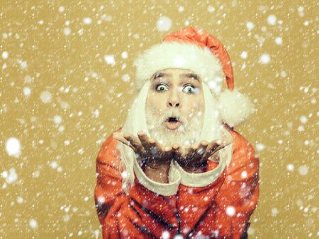 New year man with funny face with long white beard and hair in red santa claus christmas coat holding hands and blowing on yellow backgroundの写真素材
