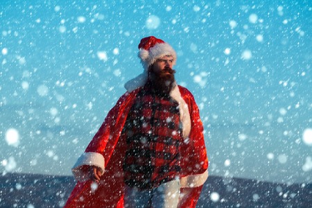 young handsome bearded bad santa claus man with long beard in checkered shirt jeans and red new year hat in christmas or xmas coat outdoor on blue sky background under white snow and snowflakesの写真素材