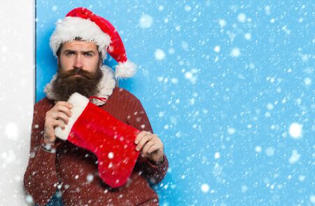young handsome bearded santa claus man with long beard in red sweater and new year hat holds...