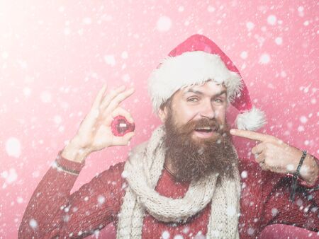 young handsome bearded santa claus man with long beard in red sweater and new year hat in scarf holds decorative christmas or xmas ball on pink studio wall background under white snow and snowflakesの写真素材