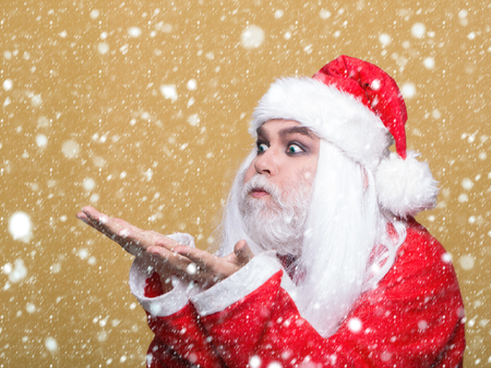 New year man with funny face with long white beard and hair in red santa claus christmas coat holding hands and blowing on yellow background under white snow and snowflakesの写真素材