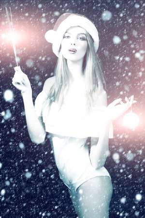 Beautiful sexual young blonde woman in white clothes hat of santa with fur and traditional new year tree ball and bengal light holding in hands standing in studio on black background, vertical pictureの写真素材