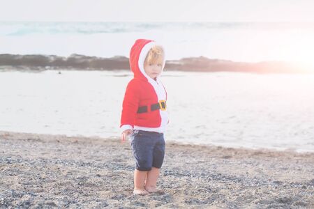 small santa claus cute boy or child barefoot in red new year coat with white fur and hat celebrates...