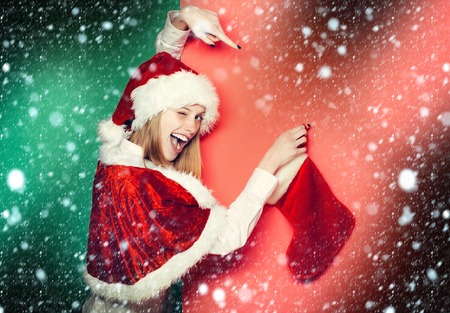 Winking pretty girl or cute beautiful woman female model in santa hat and suit with Christmas sock or stocking on pink and green background under snow and snowflakesの写真素材