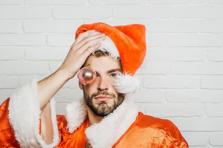 Handsome santa claus serious guy sexy young bearded macho christmas man model in red xmas or new year coat and hat on brick background holds decoration ball toyの写真素材