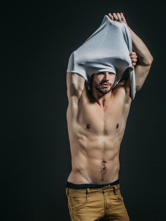 Handsome guy fashion sexy young bearded macho man model in white vest and pants has bare muscular torso and chest on grey backgroundの写真素材