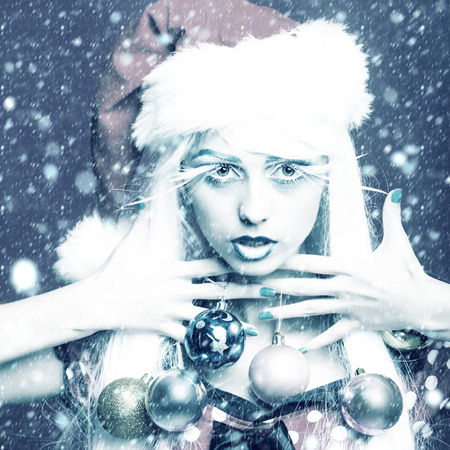 young fairy new year girl with winter makeup long white eyelashes and hair wig in christmas santa hat with decoration balls under snow and snowflakesの写真素材