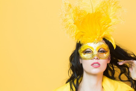 Pretty girl or cute sexy woman has brunette hair in golden festive carnival mask with feathers and yellow coat on orange background, copy spaceの写真素材