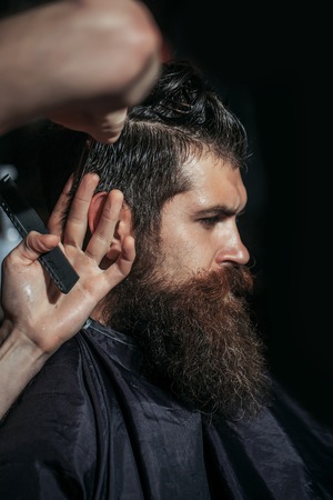 Handsome bearded man, hipster, brunette with beard and moustache has haircut in hairdressing saloon or barbershop. Barber works with scissors and combの写真素材