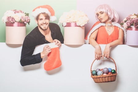young christmas couple of santa claus bearded man in red hat and pretty sexy girl in pink wig with cute smiling face near new year decoration balls in basket flowers in box and xmas stocking or sockの写真素材