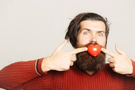 bearded amazed man in red sweater holds small heart valentines toy. handsome smiling guy with long hair isolated on white background, copy spaceの写真素材
