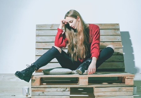 Pretty girl or beautiful woman, cute, sexy, fashion model with long, brunette, hair in casual clothes sits on wooden, pallet sofa on white wallの写真素材