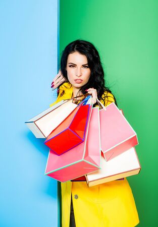 serious pretty girl or beautiful woman, young, sexy, cute, fashion model with brunette hair in yellow coat with shopping bags smiles on blue and green wall, copy spaceの写真素材