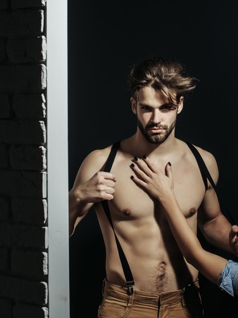 Handsome guy fashion sexy young bearded macho man model with suspenders on pants with female hand on bare muscular torso holds on brick wall and grey backgroundの写真素材