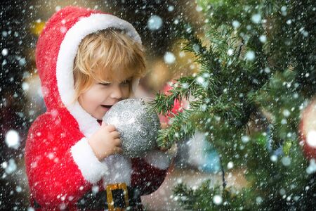 small santa claus cute boy or smiling child in red new year coat with white fur celebrates christmas or xmas holidays near green decorated tree with big balls toys under snow and snowflakesの写真素材