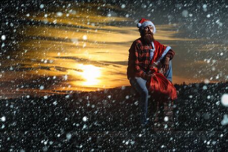 young handsome surprised bearded bad santa claus man with long beard in checkered shirt jeans and red new year hat in christmas or xmas coat with toy sack on brick chimney under snow, copy spaceの写真素材