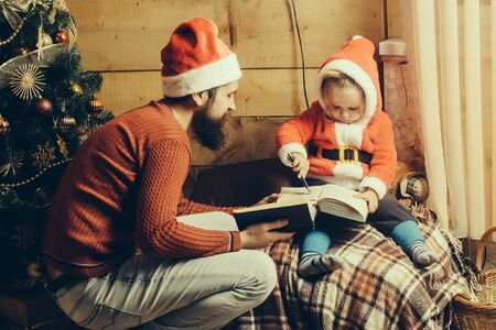 christmas father and son in red santa claus hat and sweater writing and reading book at xmas decorated tree. bearded man with cute boy child at new year holidays celebration on wooden backgroundの写真素材