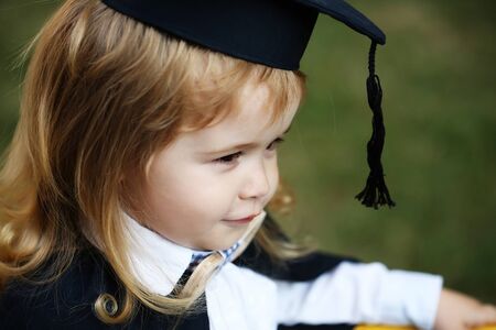 Little boy child in black academic gown and squared school hat and bow tie with blonde hair on smiling face standing outdoor on green natural backgroundの写真素材