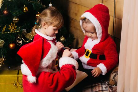 christmas happy children in red santa claus hat and coat writing and reading book at xmas decorated tree. cute kids at new year holidays celebration on wood backgroundの写真素材