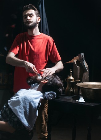 Handsome barber or shaver shaves beard to bearded man. Male customer sits in chair in barbershop or hairdressing saloonの写真素材