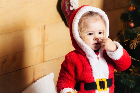 christmas small boy in red santa claus hat and coat touch nose at xmas decorated tree. cute kid at new year holidays celebration on wooden background, copy spaceの写真素材