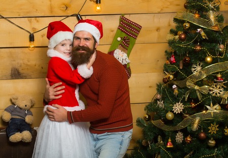 christmas father and daughter in red santa claus hat and sweater holds embraces at xmas decorated tree. bearded man with cute girl child at new year holidays celebration on wooden backgroundの写真素材