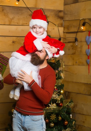 christmas father and daughter in red santa claus hat and sweater holds embraces at xmas decorated tree. bearded man with cute girl child at new year holidays celebration on wooden backgroundの写真素材