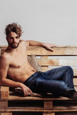 Young handsome man or muscular macho with sexy torso, sits on wooden, pallet sofa on grey background in jeansの写真素材