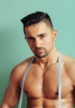 Handsome man or muscular macho, bodybuilder, with sexy, muscle torso, body, with six packs and abs poses on green background with tape measure on belly. Dieting and healthy lifestyleの写真素材
