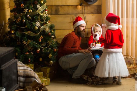 christmas father and children in red santa claus hat and sweater writing and reading book at xmas decorated tree. bearded man with cute kids at new year holidays celebration on wood backgroundの写真素材