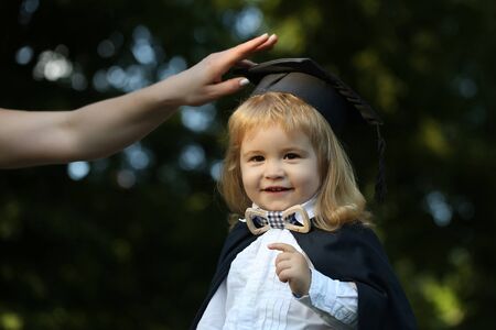 Little boy child in black academic gown and squared school hat and bow tie with blonde hair on smiling face standing outdoor on green natural background with female handの写真素材