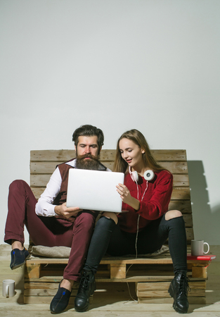 Young couple spend time together on wooden pallet sofa. Handsome man, bearded hipster with beard uses laptop, computer. Pretty girl or beautiful woman reads book, copy spaceの写真素材