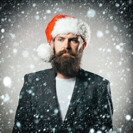 Handsome young serious man with long beard and moustache in red santa claus hat and jacket in studio...