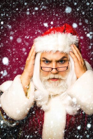 Shocked santa claus man in eyeglasses and red Christmas suit takes head in hands under snow and snowflakesの写真素材
