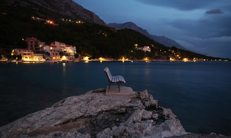 Bench on large rock in sea on blurred luminous lights coastline with houses at foot of mountains background at evening.の写真素材