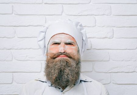 Handsome crazy man, cook or baker with flour on face, beard and moustache poses in chef uniform and...