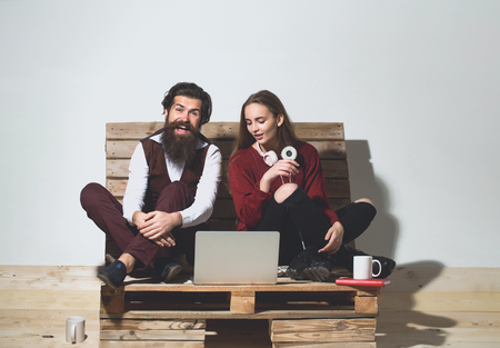 Young happy couple spend time together on wooden pallet sofa. Handsome man, bearded hipster with beard uses laptop, computer. Pretty girl or beautiful woman reads bookの写真素材