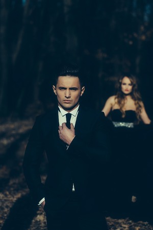 young couple of handsome man in black suit and bow with pretty sexy woman in elegant evening dress with skirt and long curly hair near tree in deep forestの写真素材