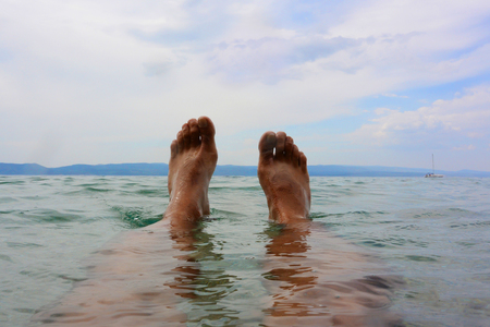 Bare male feet lie on surface of sea on cloudy sky, shoreline and floating on water yacht background.の写真素材