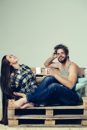 Young happy couple of pretty girl or cute woman, with brunette long hair and handsome man or muscular macho with muscular torso, sits on wooden with tea cups, pallet sofa on grey backgroundの写真素材