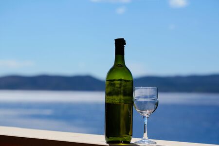 Half empty bottle and one wineglass with white wine standing on railing of balcony on blurred seascape and blue sky background.の写真素材